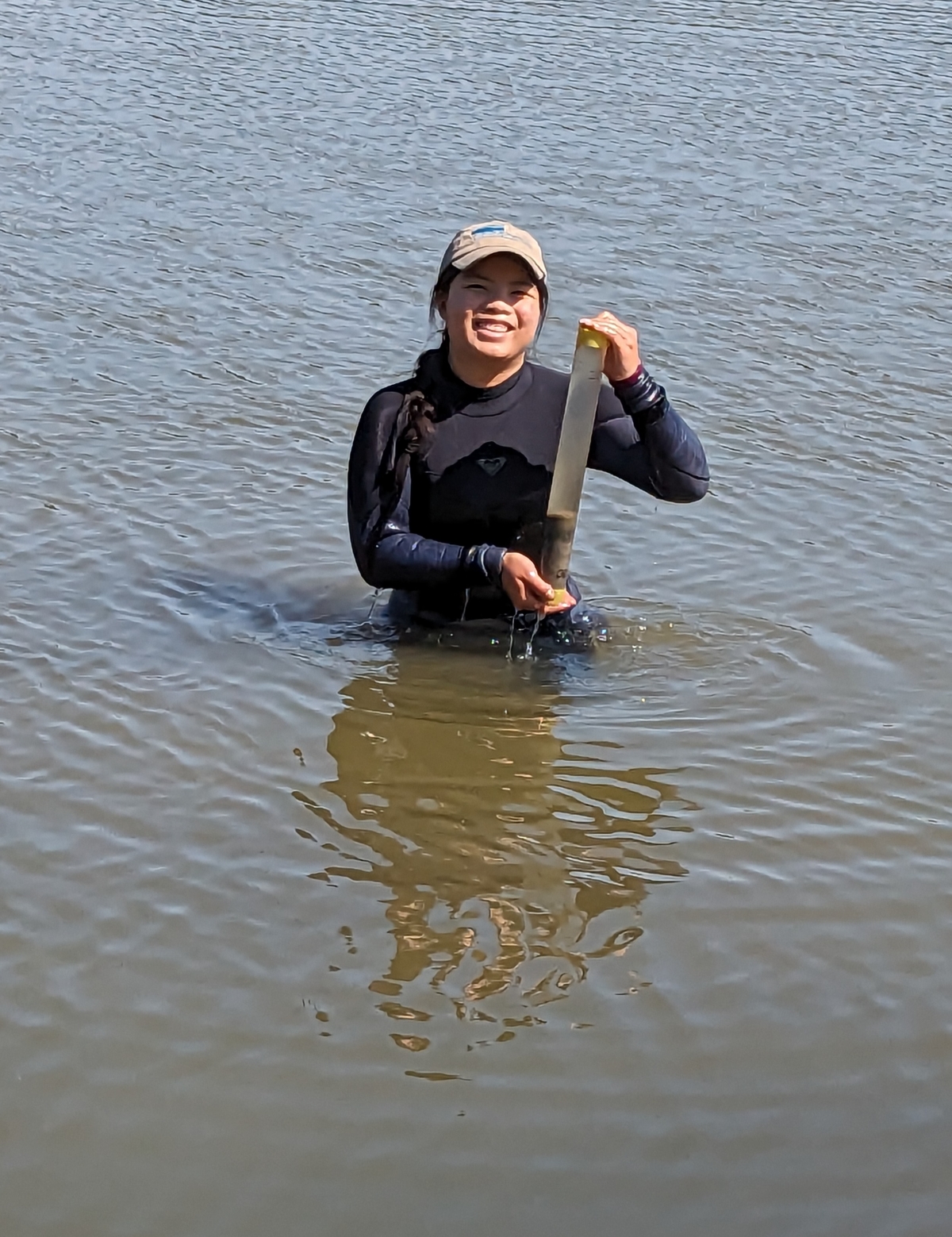 A student in a wetsuit poses with a sediment core while standing in waist-deep water.