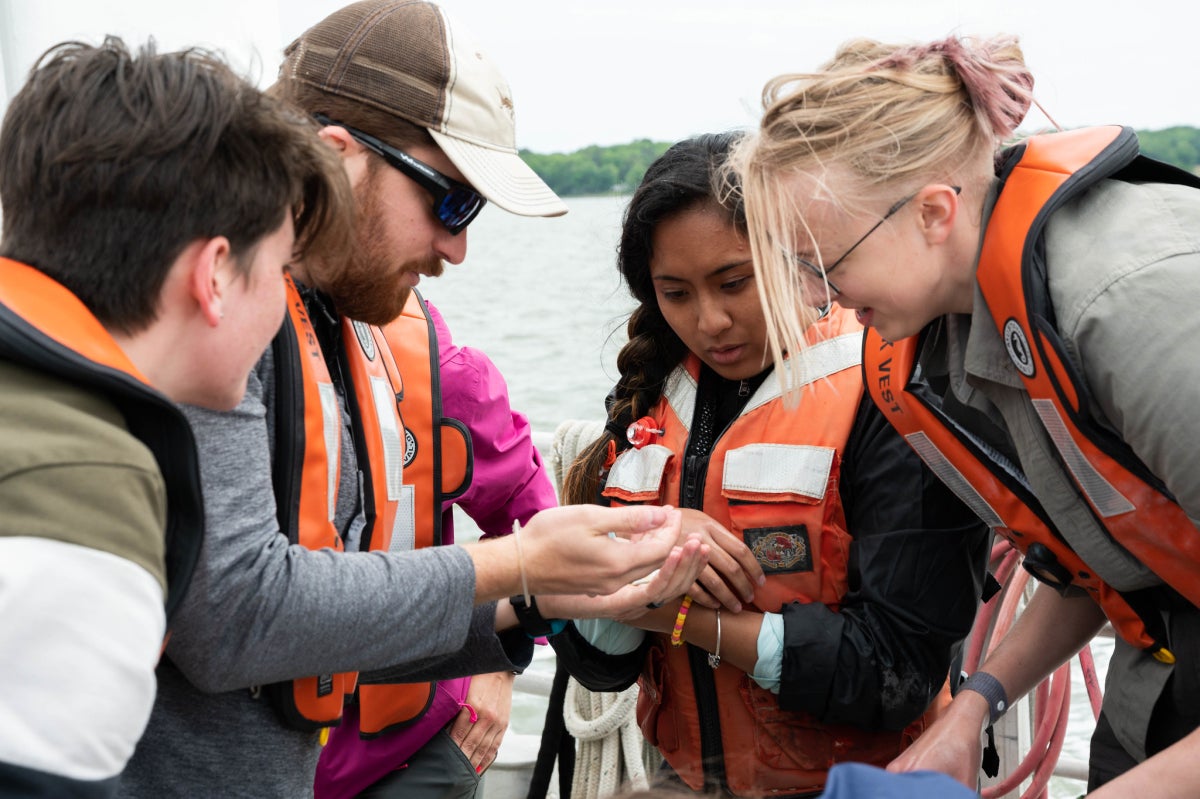 A group of students and an educator wearing life jackets aboard a boat look at something in the educator's hand