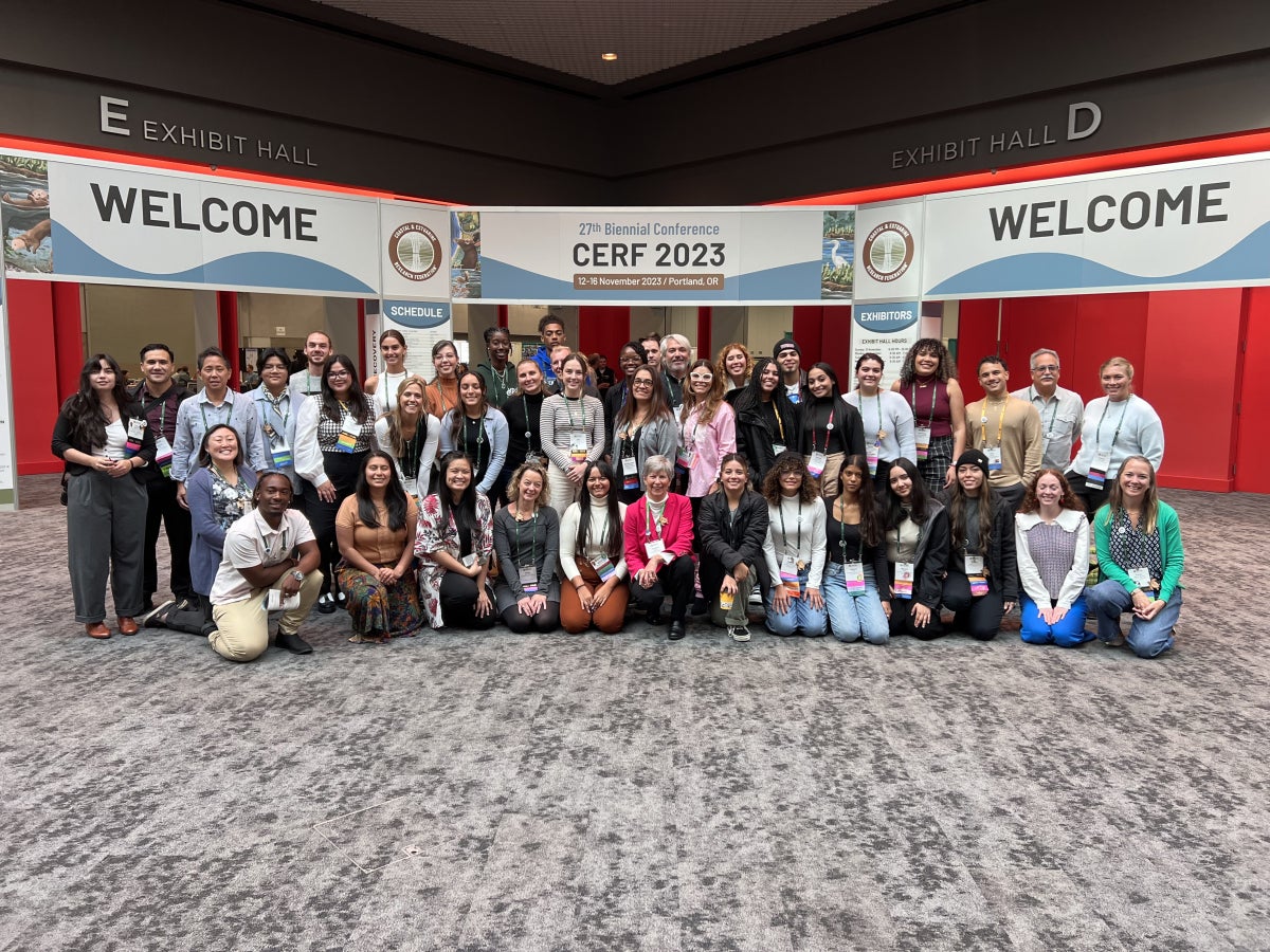 A group of about 30 students and faculty pose for a photo at the CERF 2023 conference under welcome banners