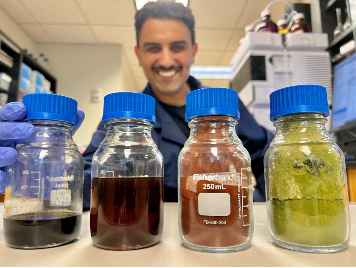 A man in a lab coat stands in the background of several bottles containing differently colored microalgae