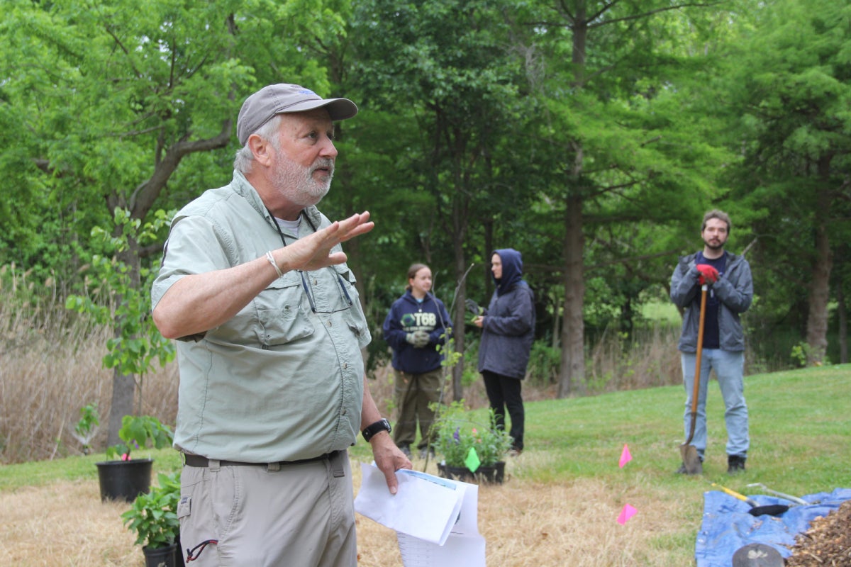 A man wearing a hat, short sleeves, and long pants holds garden planting instructions and speaks to a group of volunteers