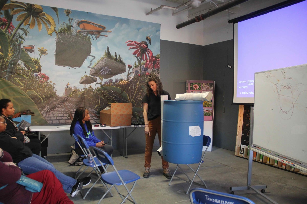 A woman stands next to a rain barrel and talks to a group of people seated in front of her