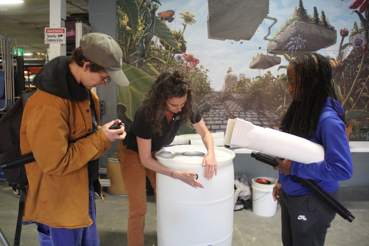 Two people watch as an instructor attaches a hose component to the top of a rain barrel